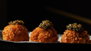 Crisp golden croquettes topped with dark caviar, in a row on a dark plate against a black background.